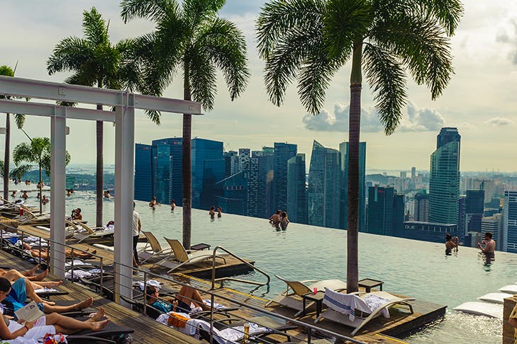 Rooftop infinity pool overlooking Singapore with people lounging by the pool under palm trees.